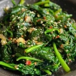 ['A close-up view of a vibrant green spinach stir fry garnished with garlic and sesame seeds.', 'A side angle shot of a healthy spinach stir fry with visible garlic cloves and red pepper flakes.', 'A detailed close-up of a colorful spinach stir fry showcasing fresh spinach and spices in a pan.', 'A mouthwatering image of sautéed spinach mixed with garlic, spices, and topped with sesame seeds.']