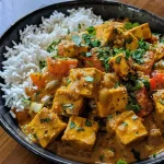 ['Close-up view of a vibrant bowl of pumpkin curry topped with crispy tofu and garnished with cilantro.', 'Side view of a colorful dish featuring pumpkin curry with golden crispy tofu, served with rice and vegetables.', 'A detailed shot of creamy pumpkin curry with chunks of tofu, tomatoes, and a sprinkle of cilantro on top.', 'Close-up perspective of a hearty pumpkin curry dish with crispy tofu and a background of naan bread.']